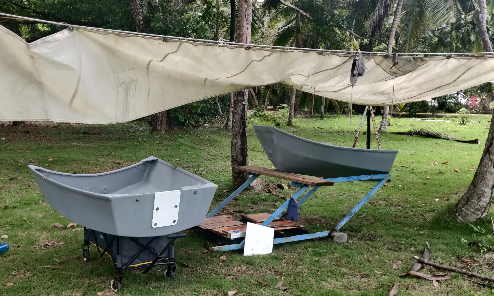 two pieces of a small boat are arranged in a makeshift workshop covered by a shade sail beneath palm trees